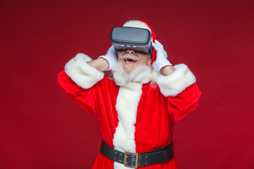Christmas. Santa Claus in black virtual reality glasses makes gestures with his hands. Surprise, emotion. New technology. Isolated on red background.