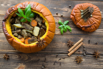 Baked meal in pumpkin on rustic brown wooden background. Stewed potatoes with carrot and roasted meat (veal and pork), decorated with basil leaves. Autumn mood, thanksgiving fall background.