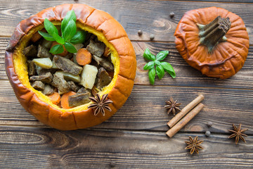 Baked meal in pumpkin on rustic brown wooden background. Stewed potatoes with carrot and roasted meat (veal and pork), decorated with basil leaves. Autumn mood, thanksgiving fall background.
