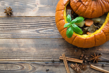 Baked meal in pumpkin on rustic brown wooden background. Stewed potatoes with carrot and roasted pork and beef, decorated with basil leaves. Autumn mood, thanksgiving fall background. Copy space.
