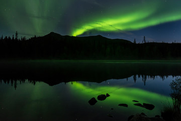Polarlicht über dem See Sjabtjakjaure mit Bergzug und Bäumen, die sich im Wasser spiegeln