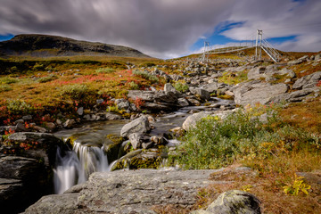 Langzeitbelichtung von Bach mit kleinem Wasserfall und Brücke auf dem Kungsleden, Lappland, Schweden