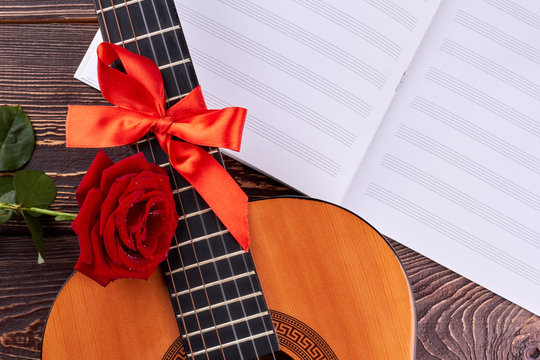 Guitar and musical notes on wooden background. Retro guitar with red ribbon. Rose and sheets of musical notes. Romantic musical background.