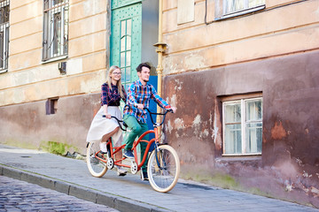 Young couple, handsome bearded man and pretty blond woman in glasses riding together tandem double red bicycle along paved sidewalk on bright sunny summer day by old buildings