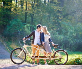 Fototapeta na wymiar Young happy romantic couple, bearded man and attractive woman close together at tandem double bicycle outdoors in summer park on blurred sunny green trees dense foliage background in the evening