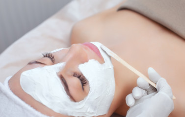 Obraz na płótnie Canvas The cosmetologist for the procedure of cleansing and moisturizing the skin, applying a mask with stick to the face of a young woman in beauty salon. Cosmetology and professional skin care.