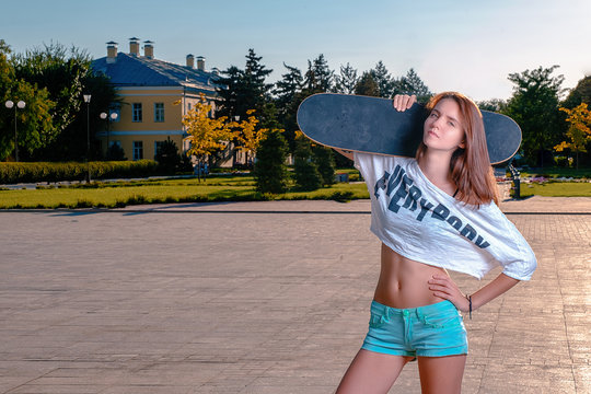 Sporty girl in short shirt showing her bare belly posing with skateboard on her shoulder, looking at camera with blank expression