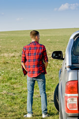 Young man in checkered shirt stand near the car on the green field