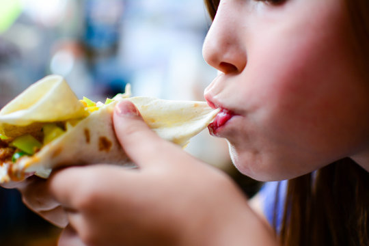 children, diet, culinary and food concept. Girl bites off tortilla with meat and vegetables. Copy space. Blurred background. 