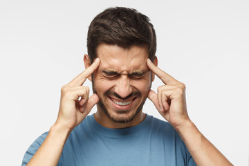 Portrait of young man isolated on grey background suffering from severe headache, pressing fingers to temples, closing eyes in order to relieve pain