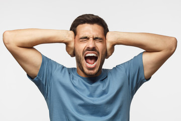 Young man in blue t-shirt, screaming with closed eyes, stressed by noise, closing ears with both...