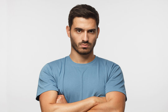 Young man in blue t-shirts tanding with arms crossed and serious concentrated face at camera, looking aggressive, isolated on gray background