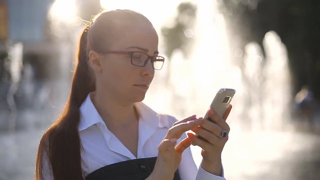 a beautiful business lady with glasses stands in the Park on the background of a fountain with a cell phone in her hand and looks at the financial news on his screen.