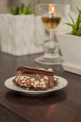 Fototapeta na wymiar Handmade raw chocolate with hazelnuts is a healthy snack and a delicious dessert. Slice of chocolate is on a decorative plate and an olive oil candle burning on a blurred background