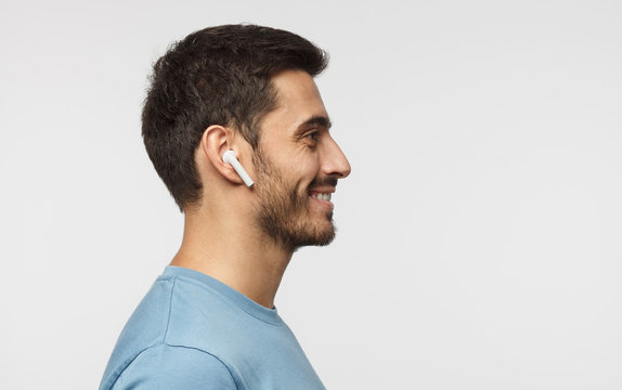 Sideways portrait of smiling young man listening to music or radio, uses modern wireless earphones, wearing blue t-shirt. Copy space for text