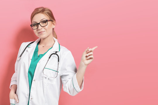 Woman doctor on pink background points something. Photo with copyspace for your text.