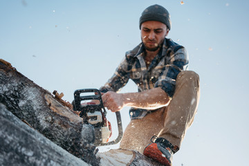 Bearded strong lumberjack wearing plaid shirt using chainsaw for work on sawmill.  Wooden chips fly...