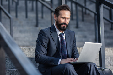 Positive bearded businessman using laptop on the staircases