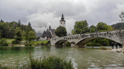Fototapeta na wymiar Church and bridge on Bohinj lake in slovenia. Very cloudy day