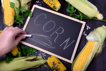 Raw and grilled maize cobs are framing the blackboard with the word "Corn" written with a piece of chalk