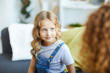 Blonde little girl in casualwear sitting on sofa at home and looking at her mom during talk