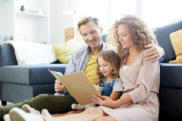 Young family of three reading book of stories while sitting on the floor by sofa at leisure