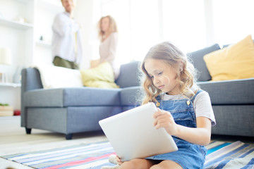 Little girl with touchpad sitting on the floor of living room and watching cartoons