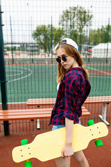 A pretty blond girl wearing checkered shirt, white cap and sunglasses is standing on the sports field with a yellow longboard in her hand. Sport and cool style.