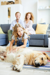 Litle girl cuddling fluffy dog lying on the floor of living-room with her parents sitting on background