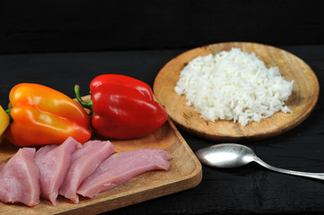 Orange and red pepper on a wooden plate. Next to pepper raw steaks from turkey meat. Bowl with boiled rice. Dark background. Close-up.