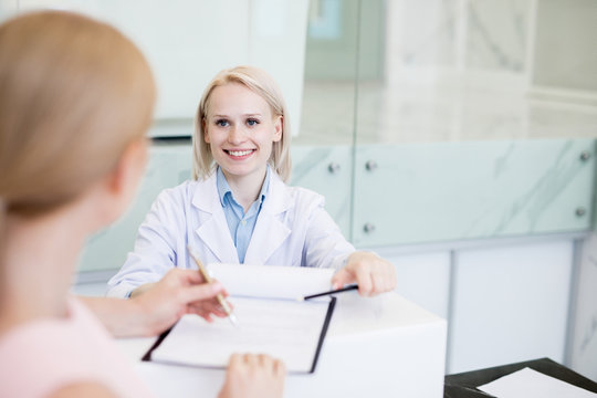 Happy young woman in whitecoat asking her patient to sign medical document during consultation