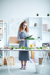 Young contemporary designer of floristics in long grey striped apron looking at you while standing by desk