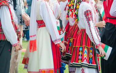 Unrecognizable irls in ethnic Bulgarian costumes with colorful ornament holding a flag of Bulgaria. Sunset at the background - obrazy, fototapety, plakaty