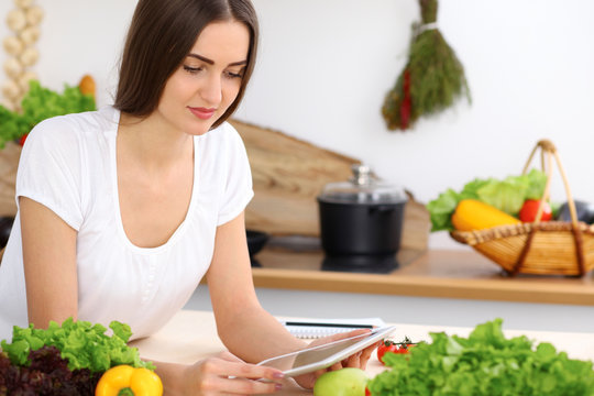 Beautiful Hispanic  Woman Cooking While Using Tablet Computer In Kitchen Or  Making Online Shopping By Touchpad And Credit Card. Housewife Found New Recipe For Dinner