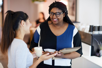 Two intercultural girls discussing ideas while one of them having drinks and her friend making notes in notepad