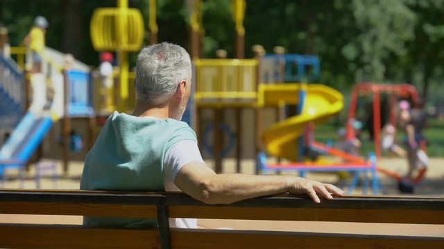 Male pensioner sitting on bench and watching grandkids on playground, family