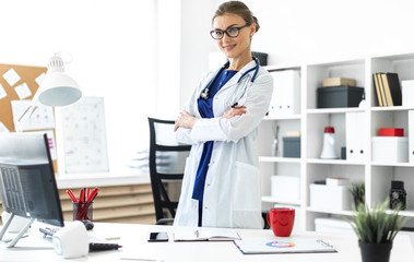A young girl in a white coat is standing near a table in her office. A stethoscope hangs around her...