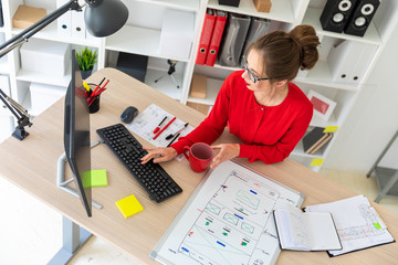 A young girl is sitting at the desk in the office, holding a red cup by her hand and typing on the...