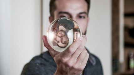 Young man holds crystal ball in front of face reflecting himself upside down with a flare of light in his reflection. - Powered by Adobe