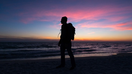 Guy stands in front of dramatic sunset over the ocean on a beach covered with waves.