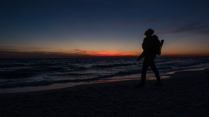 Fototapeta premium Guy stands in front of dramatic sunset over the ocean on a beach covered with waves.