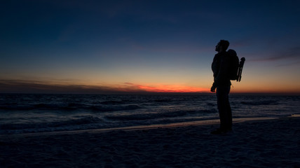 Obraz premium Guy stands in front of dramatic sunset over the ocean on a beach covered with waves.