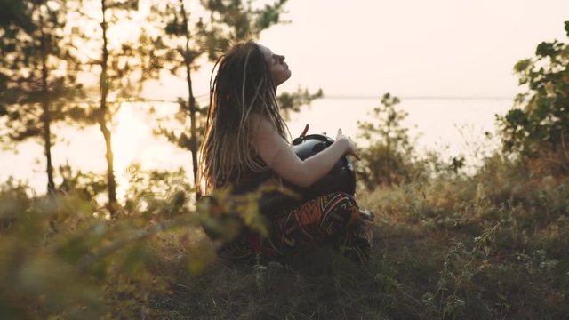 Beautiful young hippie woman with dreadlocks playing on djembe. Funky woman drumming in nature on an ethnic drum at sunset or sunrise