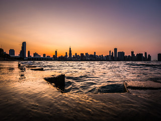 Chicago skyline picture during a beautiful sunset with purple and orange sky above and building silhouettes with rippling waves and water of Lake Michigan and concrete shoreline in the foreground.