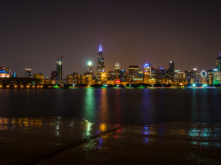 Beautiful long exposure Chicago night skyline photo with colorful red, green, purple, blue, orange, and yellow building lights and water reflections on Lake Michigan and shoreline in foreground.