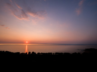 Gorgeous panoramic sunrise view over lake Michigan with colorful pink, purple and blue sky above and feathery clouds reflecting on the water below and tree lined shoreline and beach in the foreground.