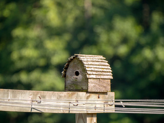 Close up photograph of a weathered wood bird house sitting on top of an outdoor wash line with blurred bokeh of trees in backg