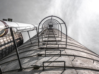 Dramatic photograph looking up the side of a concrete grain silo with metal ladder and cage and gray cloudy sky above.