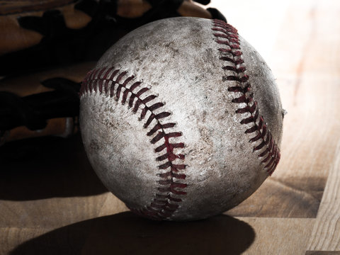 Close up sports background image of an old used weathered leather baseball laying in front of a ball glove on a wood butcher block counter showing intricate detailing and red laces.