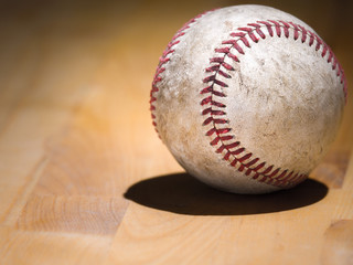 Close up sports background image of an old used weathered leather baseball showing intricate detailing and red laces sitting on a wood butcher block counter with lighting from behind and dark shadows.